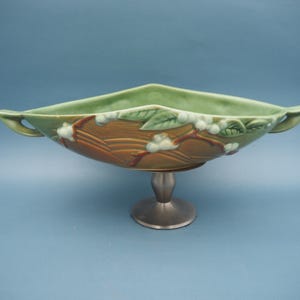 Peut inclure: Un bol décoratif en céramique avec un intérieur et un extérieur verts, orné d'un motif floral marron et blanc. Le bol a des poignées et repose sur un socle argenté. Le bol mesure environ 30 cm de long.