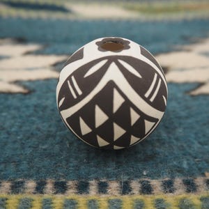 May include: A round, black and white ceramic bead with a decorative geometric pattern. The bead features a series of triangles and curved lines, and a small hole at the top. The bead is set against a blue and white patterned background.