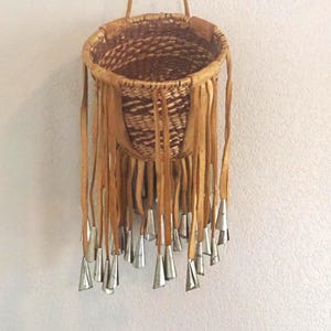 May include: A hanging woven basket featuring a tan leather fringe trim around the rim. Each leather strip is adorned with a small, conical silver-colored metal bell at the end. The basket has a circular opening and a natural brown woven texture.