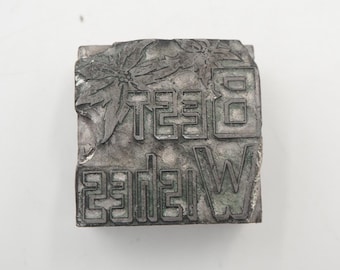 May include: A weathered, rectangular metal printing block with the words "Best Wishes" in a stylized font. The block features floral designs and is gray with hints of green.