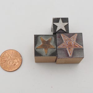 May include: A collection of vintage star-shaped printing blocks in various designs. The blocks are made of metal and wood, with copper and silver star inlays. A U.S. penny is included for size reference.