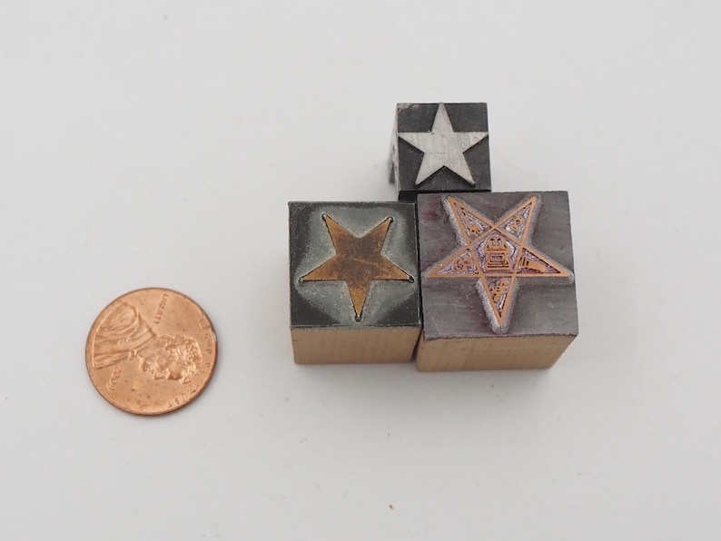 May include: A collection of vintage star-shaped printing blocks in various designs. The blocks are made of metal and wood, with copper and silver star inlays. A U.S. penny is included for size reference.