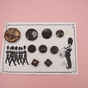 May include: A selection of vintage buttons in shades of brown and bronze, displayed on a card with an illustration of marching soldiers and a drummer. The buttons vary in shape, including round and square designs, and are of various sizes.