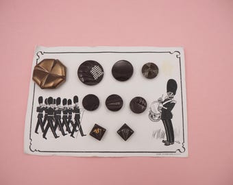 May include: A selection of vintage buttons in shades of brown and bronze, displayed on a card with an illustration of marching soldiers and a drummer. The buttons vary in shape, including round and square designs, and are of various sizes.