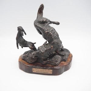May include: A bronze sculpture titled "Nip and Tuck" by Rusty Phelps. The sculpture depicts three rabbits in a dynamic pose, with one leaping upwards. The base is a dark brown colour, and the sculpture has a detailed, textured surface.