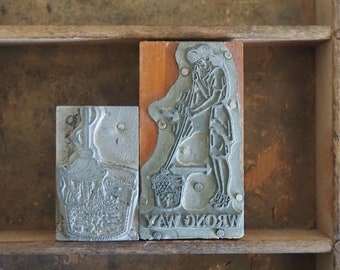 May include: Two vintage wood printing blocks with metal type. The left block shows a woman holding a broom and a bucket. The right block reads "WRONG WAY".
