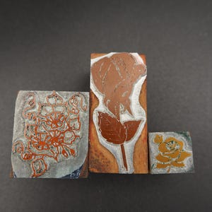 May include: Three rectangular wooden printing blocks featuring raised relief designs. The left block displays a detailed floral pattern in orange, the center block shows a single rose in brown, and the right block features a small yellow rose.