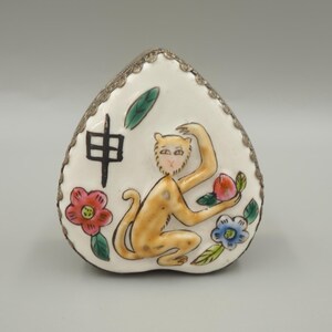 May include: A heart-shaped, white ceramic trinket box with a silver-colored trim. The box features a painted monkey holding a peach, with a black Chinese character and colorful flowers.