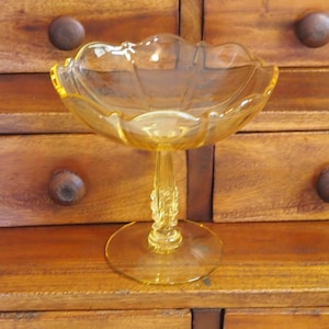 Op de afbeelding: Een vintage amberkleurige glazen voetbakschaal. De kom heeft een geschulpte rand en staat op een decoratieve steel en een ronde voet. Het glas heeft een lichte gele tint. De kom wordt waarschijnlijk gebruikt voor het serveren van fruit of snoep.