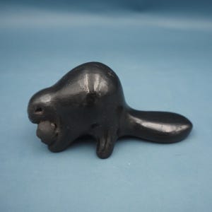 May include: A small, black ceramic beaver figurine. The beaver is depicted in a side profile, with a rounded head, small eye, and a rounded tail. The beaver is holding a small, dark object in its mouth.