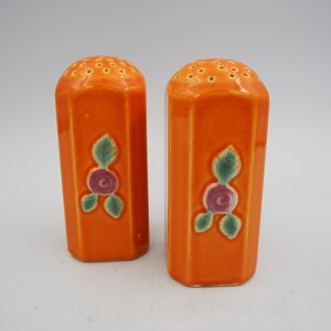 May include: A pair of bright orange ceramic salt and pepper shakers with a hexagonal shape. Each shaker features a hand-painted floral motif consisting of a small purple flower surrounded by three green leaves on the front face and a perforated top.