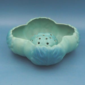 May include: A light blue ceramic flower frog with a central perforated sphere, nestled within a petal-shaped bowl. The flower frog is designed for arranging flowers and has a matte finish.