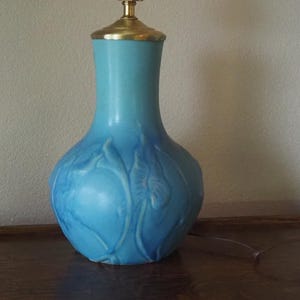 Op de afbeelding: Een turquoise keramische tafellamp met een messing hals en eindstuk. De lampvoet heeft een bolle vorm met reliëfbladmotieven. De lamp staat op een donker houten oppervlak.