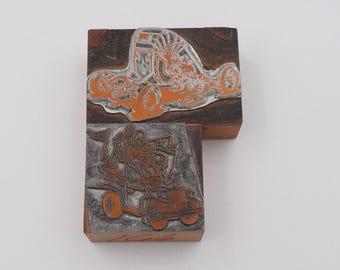 May include: Two vintage wood printing blocks with orange and silver illustrations of vehicles. The top block depicts a race car, and the bottom block shows a car with passengers. The blocks are made of dark wood.