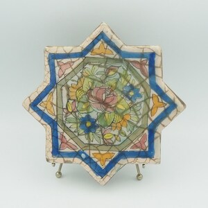May include: An eight-pointed star-shaped ceramic tile with a floral design. The tile features a central bouquet of pink, yellow, and blue flowers, surrounded by a green and blue border. The outer edge is blue with yellow accents.
