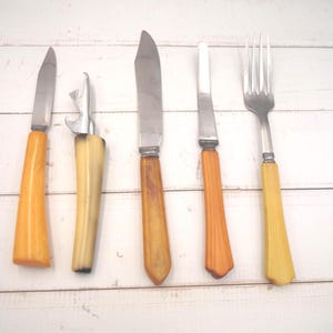 May include: A collection of vintage cutlery, including knives, a bottle opener, and a fork. The handles are in shades of yellow and orange, with stainless steel blades and opener. The items are arranged on a white wooden surface.