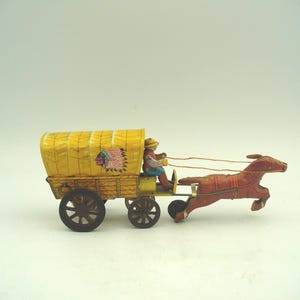 Könnte beinhalten: Ein Blechspielzeug, das einen von einem Pferd gezogenen Planwagen darstellt. Der Wagen hat ein gelbes Verdeck mit einem Indianerkopfschmuck-Motiv, einen holzfarbenen Aufbau und dunkle Räder. Eine Figur mit Hut sitzt auf dem Bock und hält die Zügel.