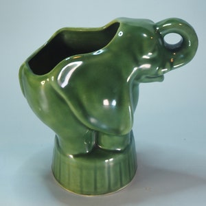 Könnte beinhalten: Ein grüner Keramik-Elefantentopf mit einem strukturierten Sockel und einem erhobenen Rüssel. Der Topf hat eine breite Öffnung oben zum Pflanzen.