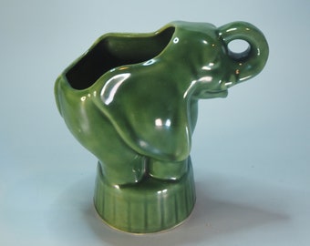 May include: A green ceramic elephant planter with a textured base and a raised trunk. The planter has a wide opening at the top for planting.