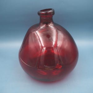 May include: A red glass bottle with a rounded body and a narrow neck. The bottle has a glossy finish and appears to be made of thick glass. The bottle's shape is reminiscent of a vintage design, suitable for decorative purposes.