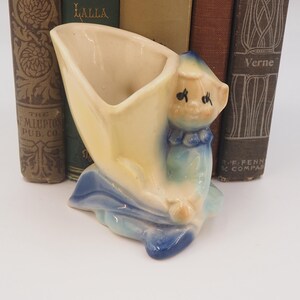May include: A ceramic planter shaped like a yellow and blue cartoon character holding a yellow flower pot. The character has a blue dress and a blue bow around its neck.