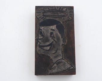 May include: A vintage woodblock print of a smiling cartoon man with a halo above his head. The print is in black and white with a brown background.