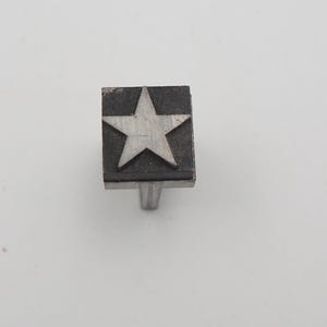 May include: A silver-toned cabinet knob with a star design. The star is centered on a square, dark-colored face. The knob has a simple, cylindrical stem for mounting. The star is a classic decorative element.