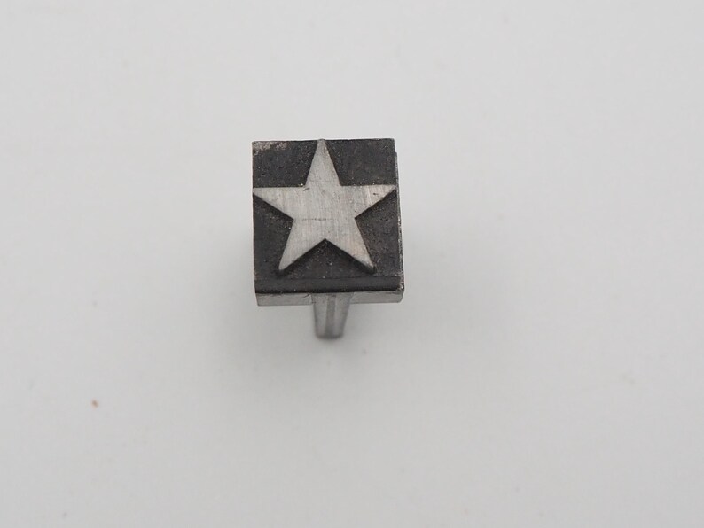 May include: A silver-toned cabinet knob with a star design. The star is centered on a square, dark-colored face. The knob has a simple, cylindrical stem for mounting. The star is a classic decorative element.