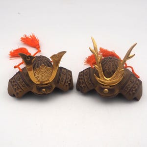 May include: Two miniature samurai helmets, known as kabuto, featuring dark brown textured bowls and gold-coloured metal crests. Each helmet is adorned with bright orange tassels attached to the back, showcasing intricate detailing on the front plates.