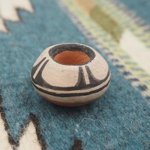 May include: A small, round, hand-painted ceramic pot with a wide opening. The pot is primarily off-white with black and brown geometric designs. The rim is black, and the interior is a warm brown color. The pot is likely a decorative item or a small container.