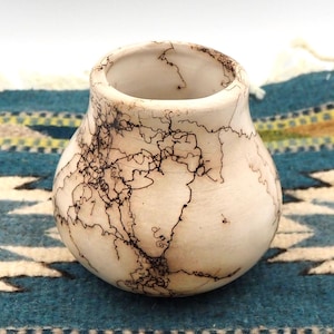 May include: A cream-coloured ceramic vase with a wide body and narrow opening. The vase is decorated with intricate black lightning-like patterns. The vase sits on a blue and white patterned textile.