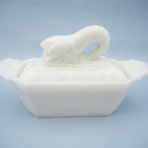 May include: A white milk glass butter dish featuring a rectangular base with scalloped edges and a lid topped with a sculpted lobster handle. The lid is decorated with a subtle embossed leaf pattern and a beaded border along the rim.