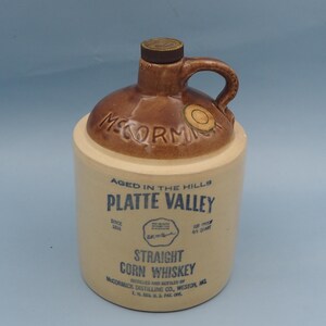 May include: A beige ceramic whiskey jug with a brown glazed top and handle. The text reads: McCormick, Aged in the Hills, Platte Valley, Straight Corn Whiskey, Distilled and Bottled by McCormick Distilling Co., Weston, MO, 4/5 Quart.