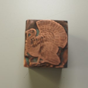 K&ouml;nnte beinhalten: Ein kupferfarbener Metallstempel mit einem detaillierten Design eines Truthahns. Der Truthahn ist nach links gerichtet und hat seine Fl&uuml;gel ausgebreitet.
