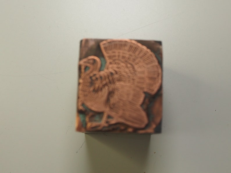 K&ouml;nnte beinhalten: Ein kupferfarbener Metallstempel mit einem detaillierten Design eines Truthahns. Der Truthahn ist nach links gerichtet und hat seine Fl&uuml;gel ausgebreitet.