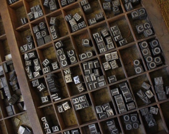 Könnte beinhalten: Ein antiker Holz-Druckkasten gefüllt mit metallenen Lettern. Der Kasten ist in Fächer unterteilt, die jeweils einen anderen Buchstaben oder ein anderes Symbol enthalten. Die Buchstaben sind alphabetisch geordnet, einige Buchstaben fehlen. Der Kasten ist aus dunklem Holz und hat ein abgenutztes, antikes Aussehen.