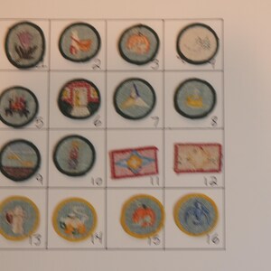 Op de afbeelding: Een wit vel papier met 16 ronde plekken, elk met een ander ontwerp, erop genaaid. De plekken zijn genummerd van 1 tot en met 16. De plekken zijn allemaal in verschillende kleuren en ontwerpen, waaronder een pompoen, een huis, een vogel, een bloem en een zeilboot.