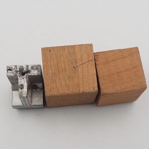 May include: An assortment of small wooden and metal blocks. Two square wooden blocks are side by side, with a third, smaller, metal block to the left. The wood is a light brown color, and the metal block is silver.