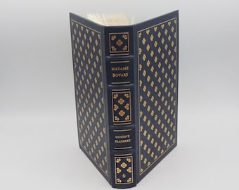 May include: A hardcover book titled "Madame Bovary" by Gustave Flaubert. The book has a navy blue cover with a gold floral pattern and gold lettering on the spine. The pages are white.
