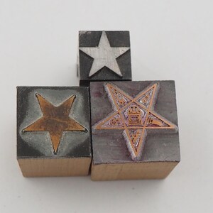 May include: Three vintage printing blocks with star designs. One block features a silver star, another a copper-colored star, and the third a detailed pentagram design. The blocks are made of wood and metal, showcasing a mix of textures and colors.