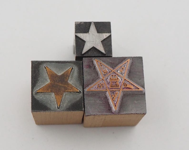 May include: Three vintage printing blocks with star designs. One block features a silver star, another a copper-colored star, and the third a detailed pentagram design. The blocks are made of wood and metal, showcasing a mix of textures and colors.