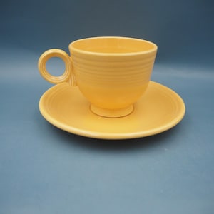 May include: A yellow ceramic teacup and saucer set. The cup has a rounded base and a circular handle. The saucer is round with a slightly raised edge. The set is a solid, bright yellow color and sits on a blue background.