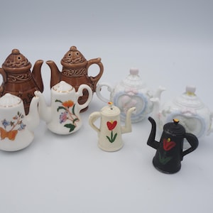 May include: A collection of ceramic teapot-shaped salt and pepper shakers. The shakers are in various colors, including brown, white, and black, and feature floral and heart designs. Some have lids, and all have spouts and handles.