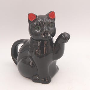 Può includere: Una teiera in ceramica nera lucida a forma di gatto seduto. Il gatto presenta orecchie interne rosso brillante, baffi dipinti in bianco e occhi verdi. Il manico è formato dalla coda del gatto, mentre il beccuccio è integrato nella zampa anteriore sollevata.