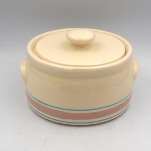 May include: A cream-colored ceramic casserole dish with a matching lid and two side handles. The base of the round pot features a wide horizontal band in dusty rose, flanked by two thin, parallel teal blue stripes that wrap around the exterior.