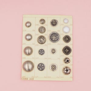 May include: A collection of vintage buttons displayed on a cream-colored card with a grid pattern. The buttons vary in size, shape, and material, including metal and pearl. The card is labeled "CARD 339" and is set against a pink background.