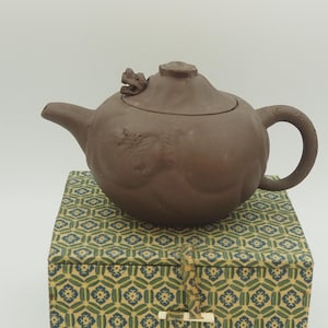 以下が含まれることがあります： 龍の形をした取っ手と蓋のつまみが付いた茶色のセラミックティーポット。 ティーポットは、緑と黄色の幾何学模様の装飾ボックスの上に置かれています。 ティーポットは丸みを帯びたボディと湾曲した注ぎ口を備えています。
