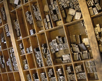 May include: A wooden printing type case filled with metal letterpress type. The case is divided into compartments, each containing a different letter or number. The type is arranged in rows and columns, and the case is made of wood with a dark brown finish.