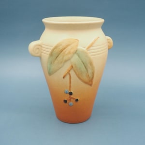 May include: A cream-colored ceramic vase with a gradient orange base. The vase features two side handles and a decorative leaf and berry design in shades of green, brown, and blue. The vase is a decorative home accent.