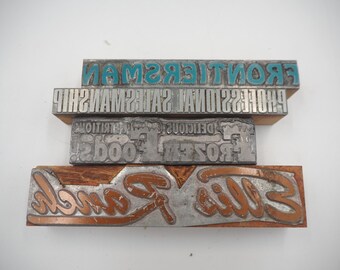 May include: A collection of vintage metal printing blocks. The blocks feature text in various fonts and sizes, including "FRONTIERSMAN", "PROFESSIONAL SALESMANSHIP", "DELICIOUS FOODS", and "Sills Foods".